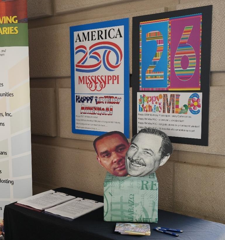 Table with notebooks and pens in front of posters reading ‘America 250 Mississippi’ and ‘Happy Birthday MLC 26’ on a stone wall.