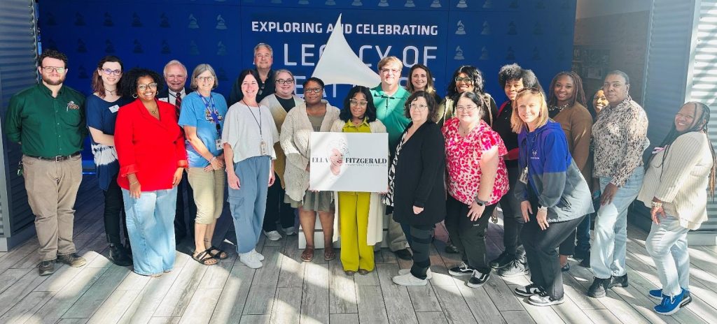 Group of people standing together indoors in front of a blue backdrop with text about exploring and celebrating legacy, holding a sign that reads ‘Ella Fitzgerald.’