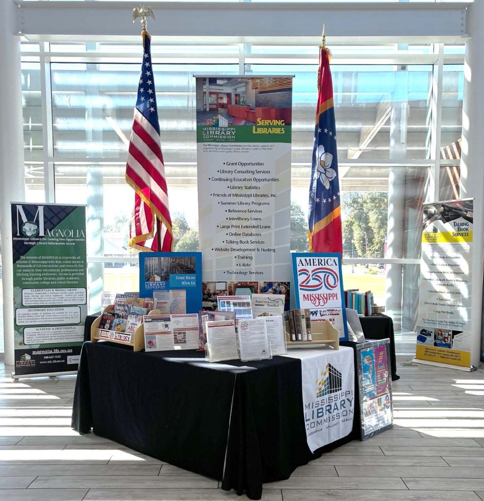 Mississippi Library Commission booth with America 250 signage, U.S. and state flags, books, and informational banners displayed in a bright glass-walled space.