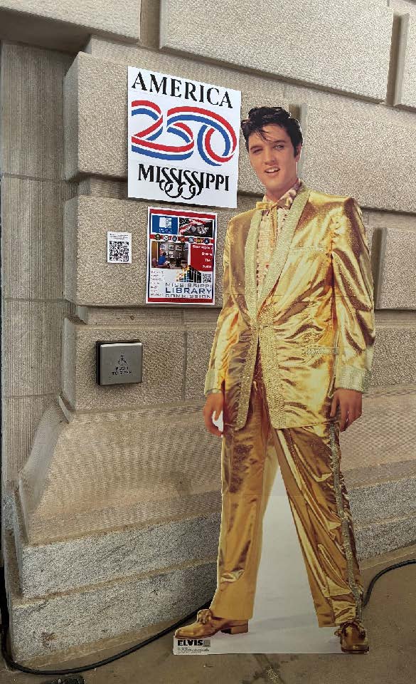 Cardboard cutout of Elvis in a gold suit next to America 250 Mississippi posters on a stone wall.