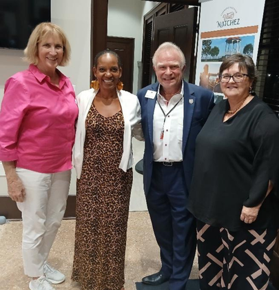 Four people standing together indoors near a banner that reads ‘Visit Natchez,’ with dark wooden doors and framed photos in the background.