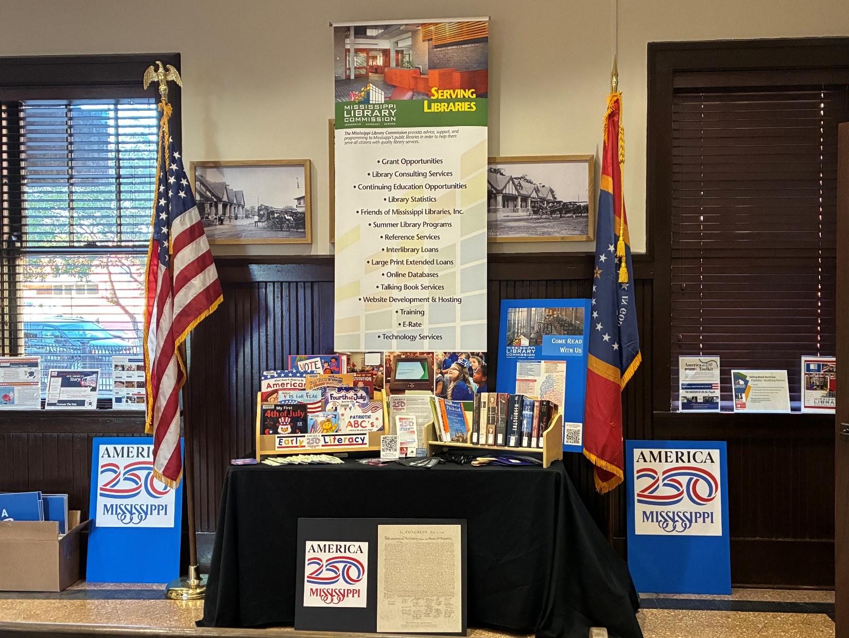 Mississippi Library Commission booth with books, America 250 posters, U.S. and state flags, and a banner listing library services.