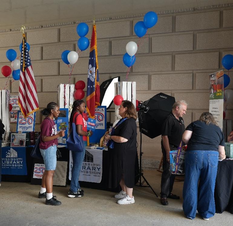People interacting at a Mississippi Library Commission booth decorated with balloons, flags, and America 250 posters.