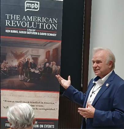 Person gesturing toward a banner for the film ‘The American Revolution’ by Ken Burns, Sarah Botstein, and David Schmidt, featuring a historical painting and text about the event.