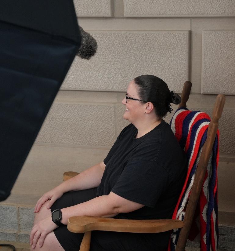 Person seated in a wooden chair with a red, white, and blue striped blanket, next to a wall and photography equipment.