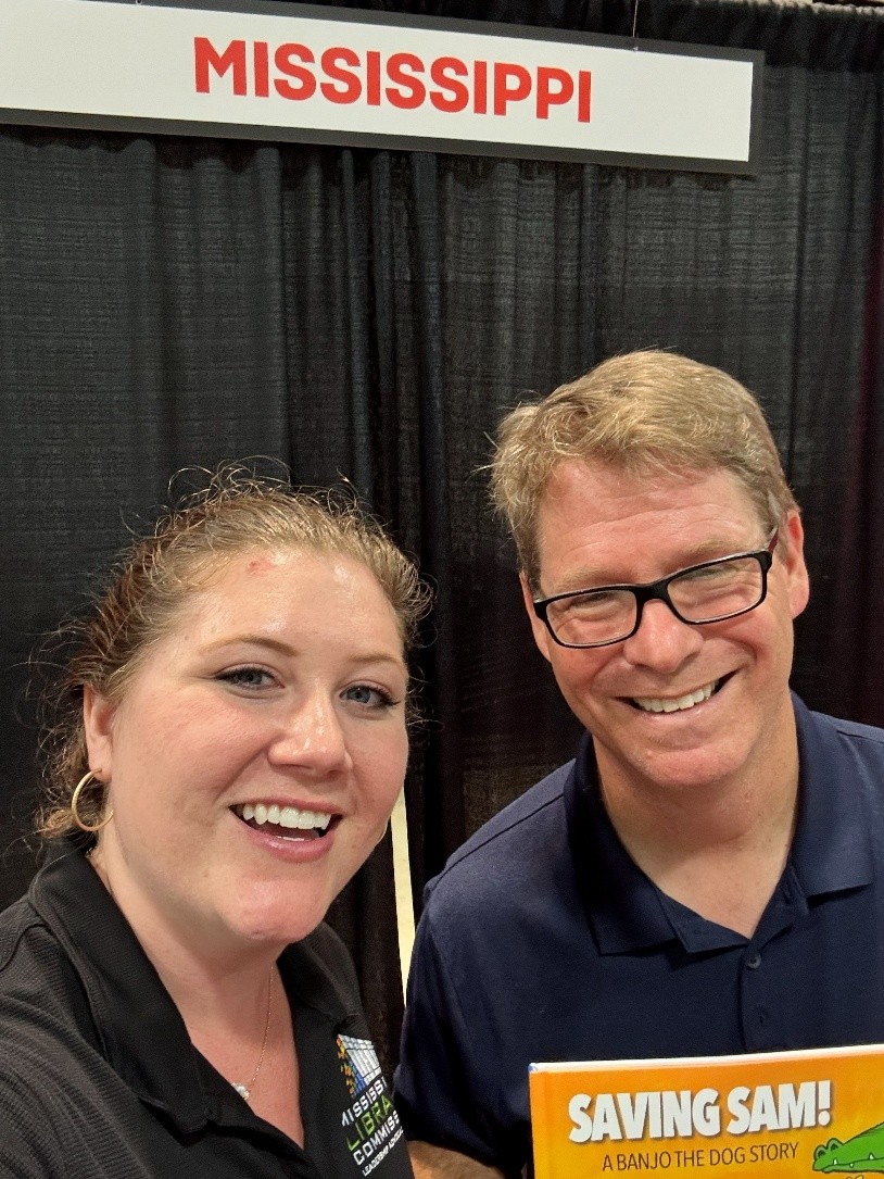 Marshall Ramsey and Alex standing in front of a black backdrop with a sign reading ‘Mississippi,’ holding a children’s book titled ‘Saving Sam: A Banjo the Dog Story.’