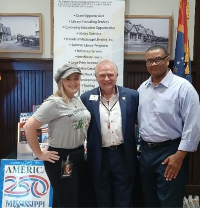 Three people standing in front of a Mississippi Library Commission booth with an America 250 poster and a banner listing library services.