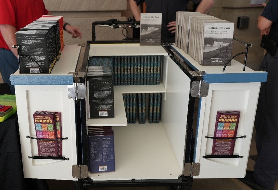 Open book bike display with shelves holding multiple books and pamphlets, including titles like ‘Cotton Gin Port’ and colorful reading guides.