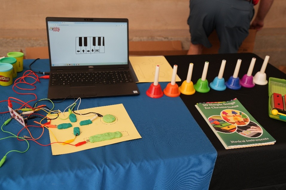 Table with a laptop showing piano keys, colorful handbells, Play-Doh containers, wires connected to a circuit board, and a booklet titled ‘36 Simple Songs for Chromaharp.’