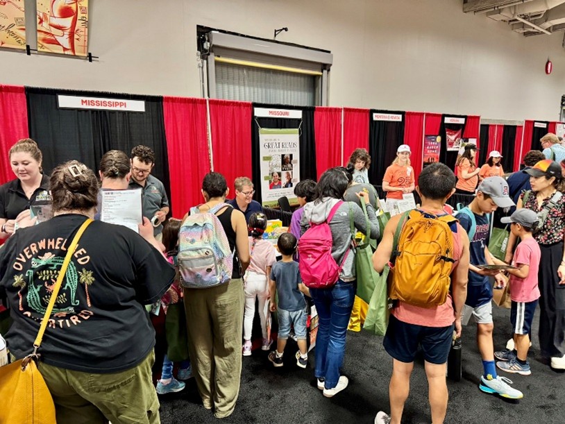 Event booth with people browsing magazines and colorful children’s books on a table, with red and black curtains and other booths visible in the background.