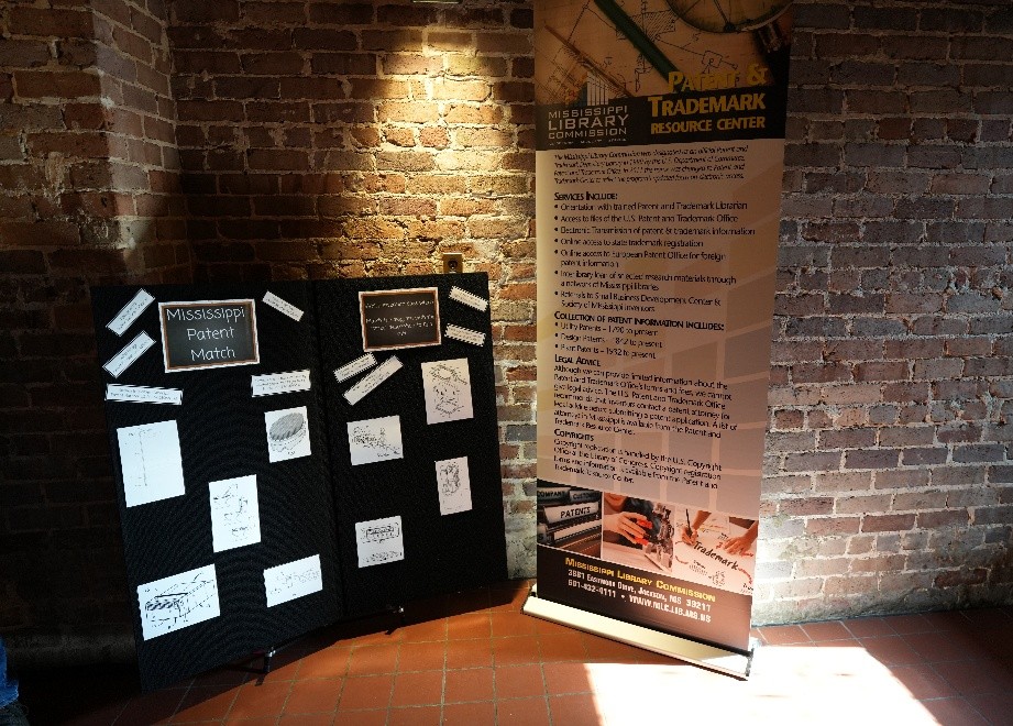 Display featuring Mississippi Patent Match boards with sketches and a banner for the Patent and Trademark Resource Center against a brick wall.
