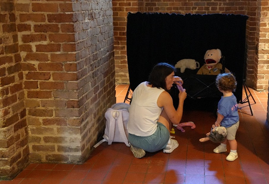 Child standing near a seated adult in front of a small puppet stage with stuffed animals and a large puppet inside a brick-walled room.