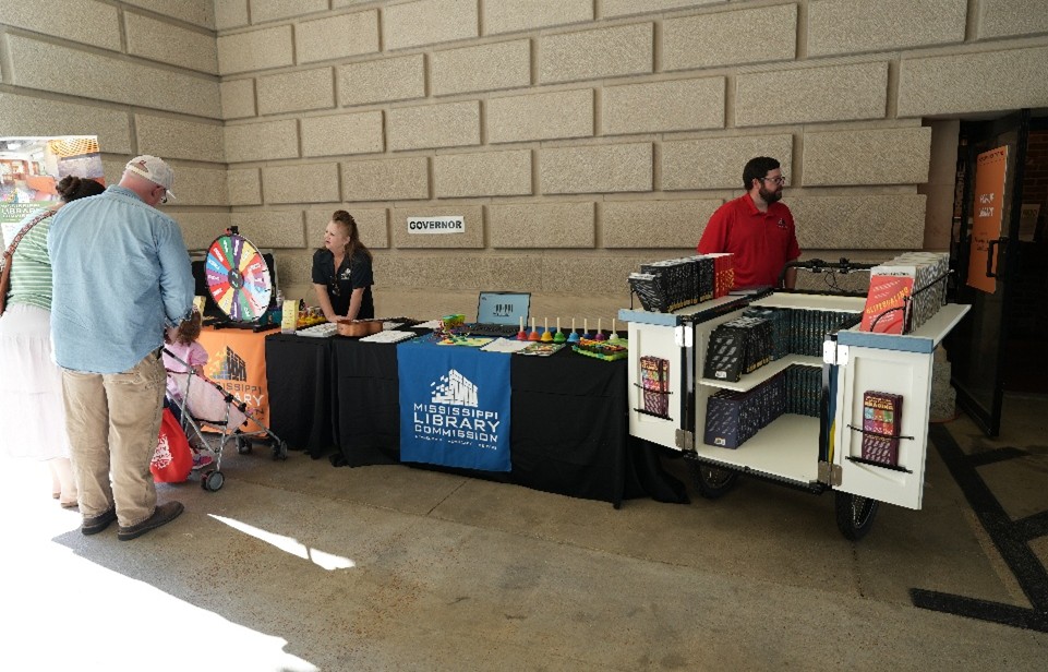 Mississippi Library Commission table with a prize wheel, colorful giveaways, and a cart filled with books in a hallway.