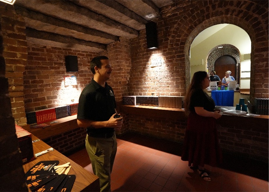 Brick-walled room with wooden beams, books on shelves, and two people standing near a counter with a window opening to another area.