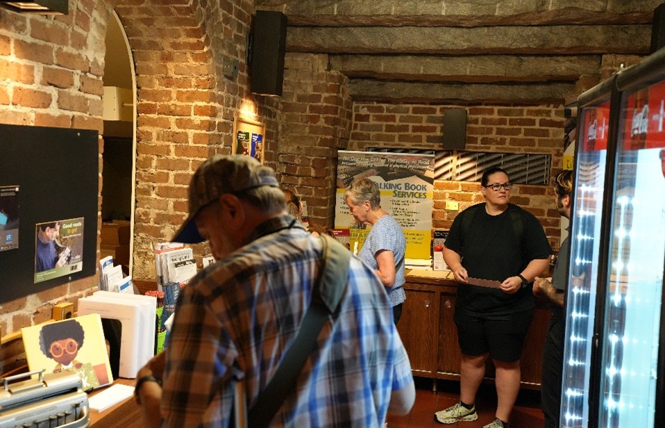 Brick-walled room with informational displays and a banner for Talking Book Services, with people browsing materials on a wooden counter.