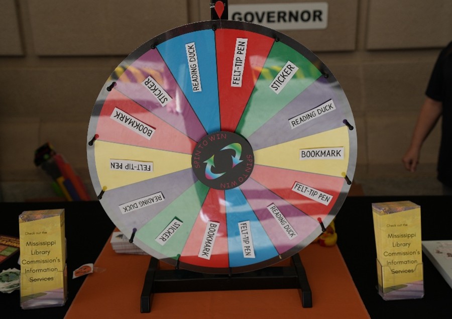 Colorful prize wheel labeled ‘Spin to Win’ with sections for items like stickers, bookmarks, reading ducks, and felt-tip pens, displayed on a table with brochures for Mississippi Library Commission’s Information Services.