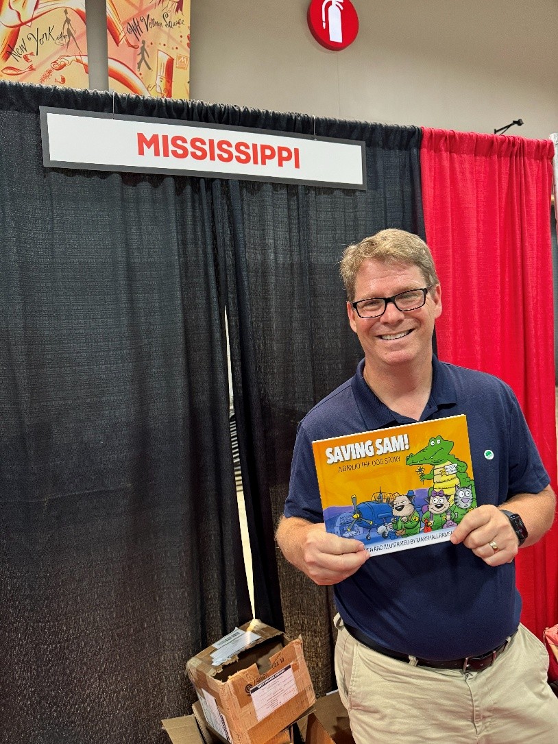 Marshall Ramsey standing in front of a black backdrop with a sign reading ‘Mississippi,’ holding a children’s book titled ‘Saving Sam: A Banjo the Dog Story,’ with boxes on the floor nearby.