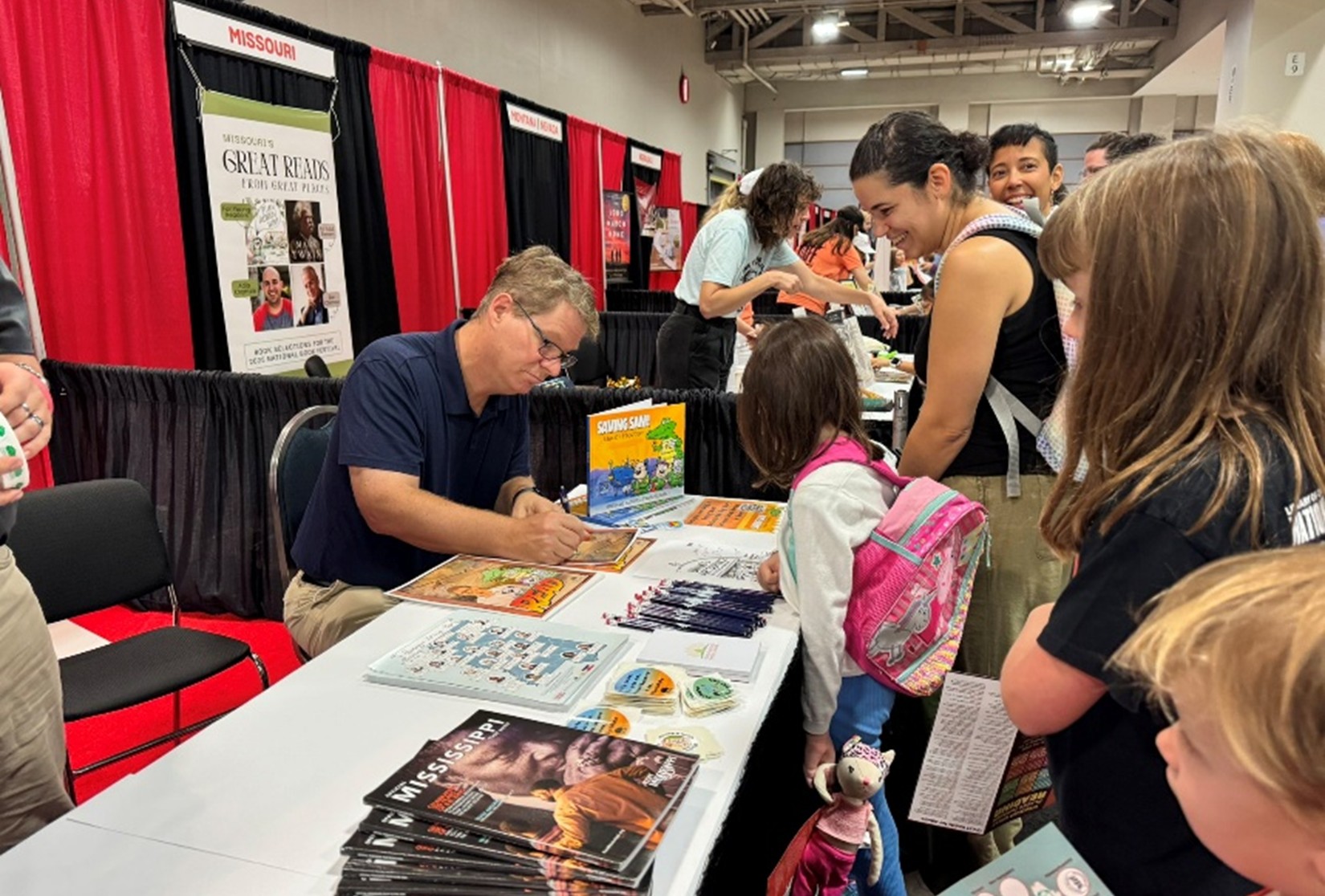 Person signing books at a table covered with magazines, brochures, and colorful children’s books, while attendees wait in line at an indoor event.