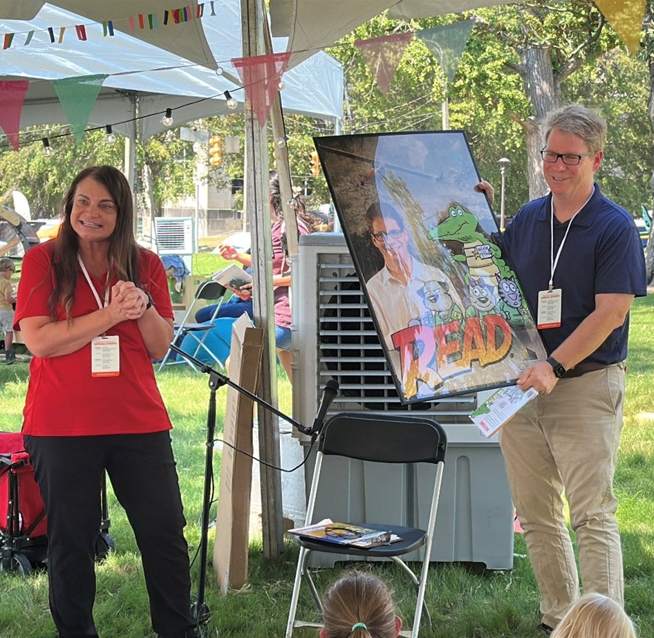 Marshall Ramsey holding a large illustrated poster with the word ‘READ’ under a tent decorated with colorful pennant banners during an outdoor event.