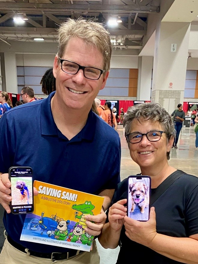 Two people holding smartphones showing dog photos and a children’s book titled ‘Saving Sam: A Banjo the Dog Story’ at an indoor event.