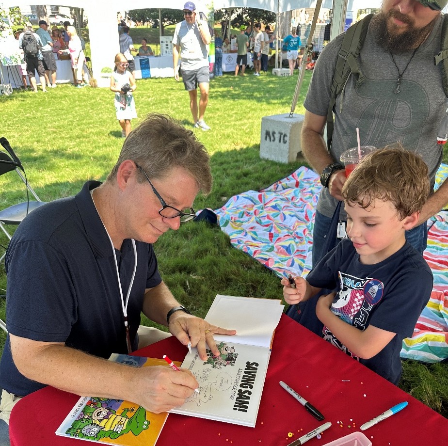 Marshall Ramsey signing a book titled ‘Saving Sam’ at a table outdoors, with markers and a colorful blanket in the background.