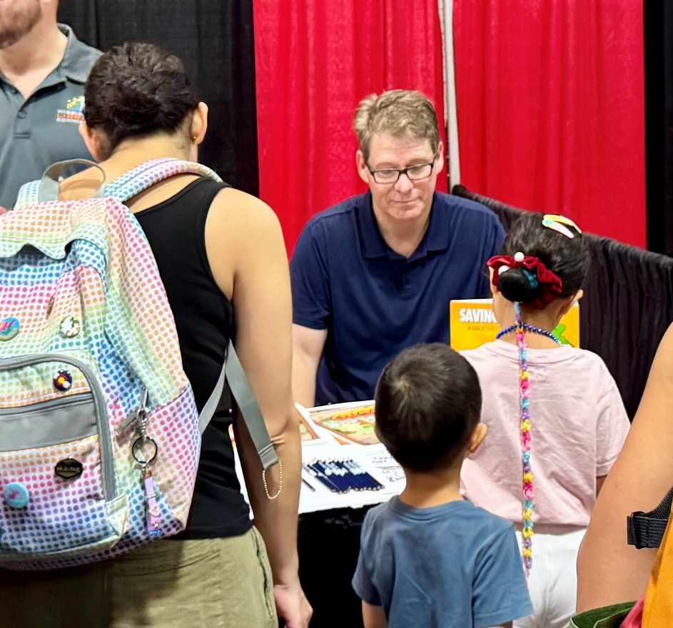 Marshall Ramsey seated at a booth with a book titled ‘Saving Sam’ on the table, interacting with children and adults standing nearby.