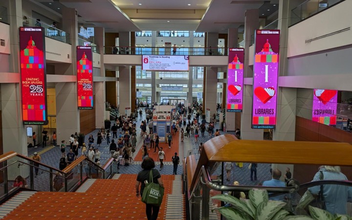 Large convention center lobby with red carpeted stairs, digital banners reading ‘I love Libraries’ and ‘Uniting Book Lovers for 25 Years,’ and crowds walking through the space.