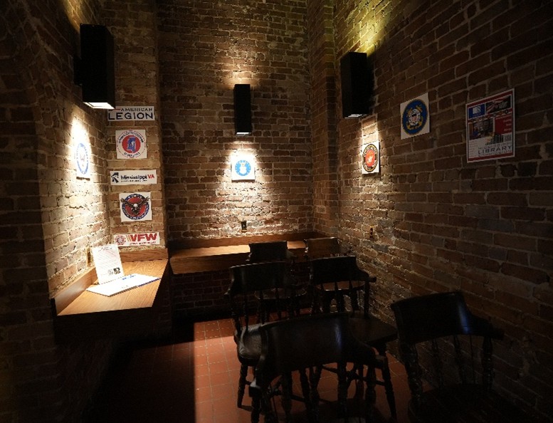 Brick-walled room with wooden chairs and tables, illuminated by wall lights, featuring posters for American Legion, VFW, and other veteran organizations.