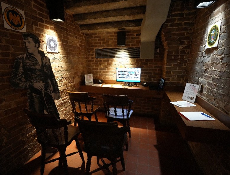 Brick-walled room with a computer on a wooden desk, chairs arranged in front, and posters for veteran organizations on the walls, with a cardboard cutout of Elvis.