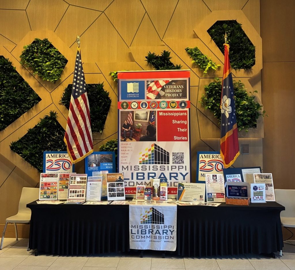 A Mississippi Library Commission display table is set up with brochures, books, banners, and two flags behind it. The center banner highlights the Veterans History Project and Mississippi library services.