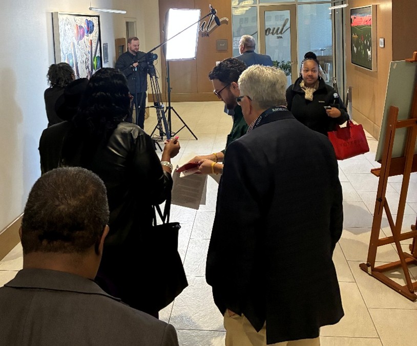 A group of attendees waits in a hallway near a video recording setup with lights and a camera operator. People are standing in line holding papers and moving toward the recording area.