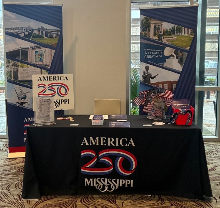 A table featuring America 250 Mississippi promotional materials is displayed with banners and patriotic décor. Brochures, flyers, and small American flags are arranged neatly across the table.