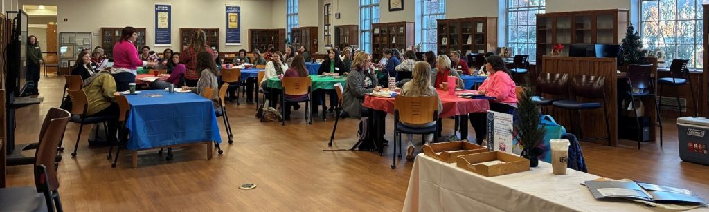 A large group of attendees sits at tables with colorful tablecloths during a session inside a library-style room. Two presenters stand at the front addressing the audience while bookshelves and large windows line the walls.