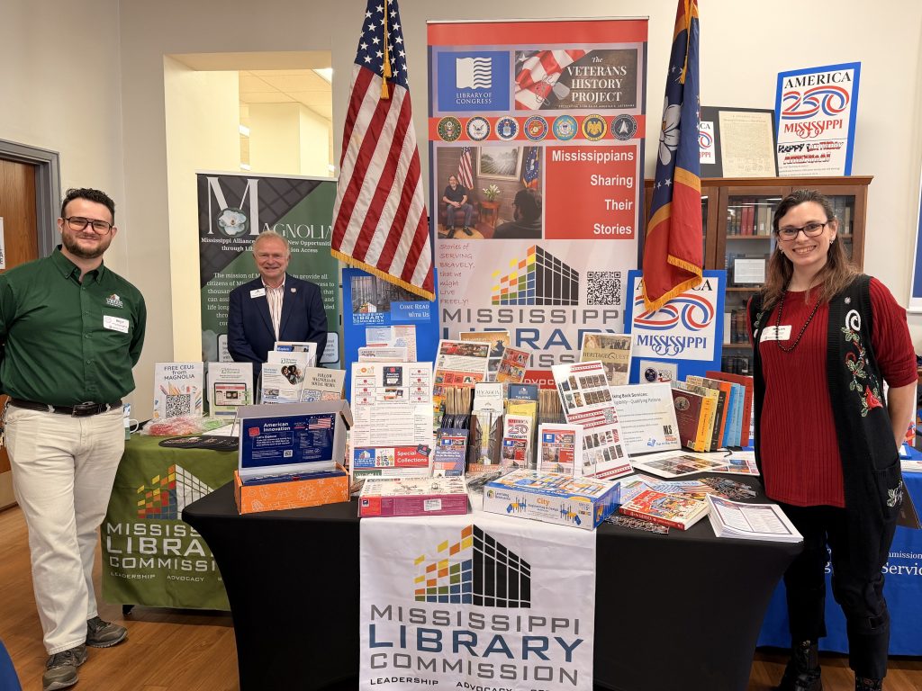Three people stand beside a large Mississippi Library Commission display table filled with books, flyers, and promotional materials. U.S. and state flags frame the colorful exhibit in the background.