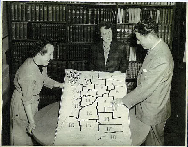 Black and white photo of three people gathered around a table examining a map labeled "Proposed Library Regions," with bookshelves in the background.