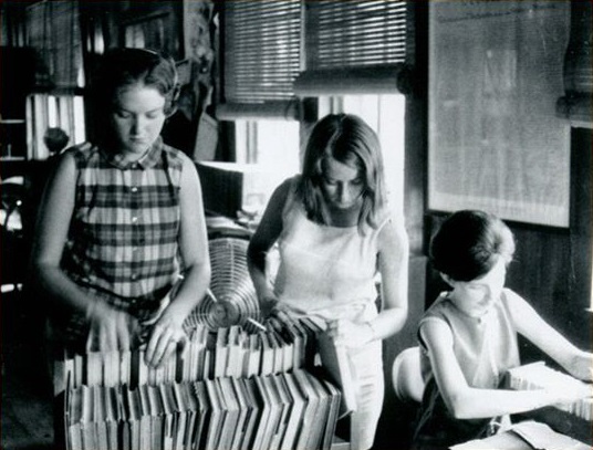Black and white photo of three women browsing and organizing books on shelves inside a library.