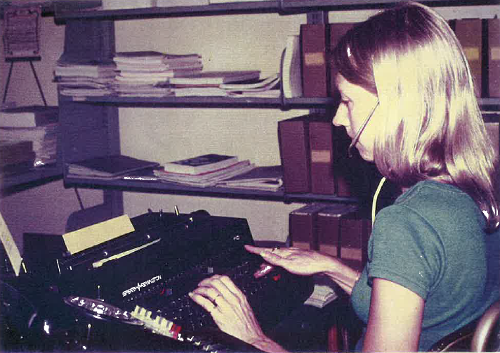 Photo of a person wearing a headset operating a telephone switchboard or communication system in an office with shelves of books and files behind.