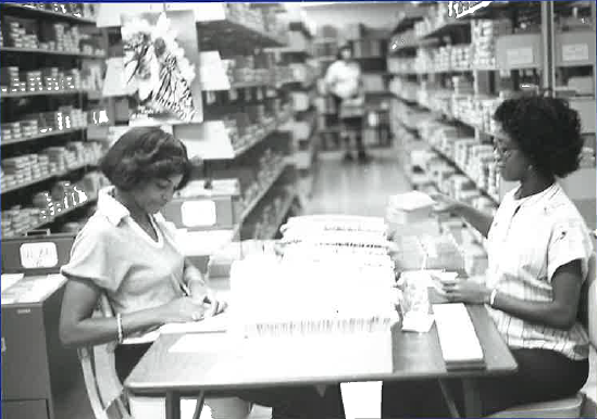 Black and white photo of two people seated at a table in a library processing materials, with shelves of items lining both sides of the room.
